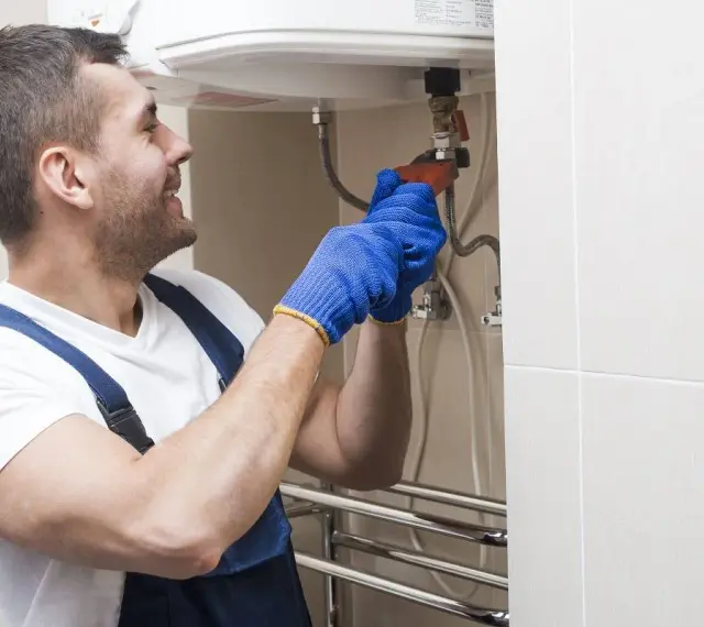 Instalator w niebieskich rękawiczkach dokręca zawór, pokazując, jak spuścić wodę z bojlera.
